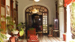 Un vestíbulo amplio con techo alto, columnas de madera ornamentadas y un gran arco de entrada a otra estancia. Hay varias sillas y mesas dispuestas para sentarse, y macetas con plantas decoran el espacio.