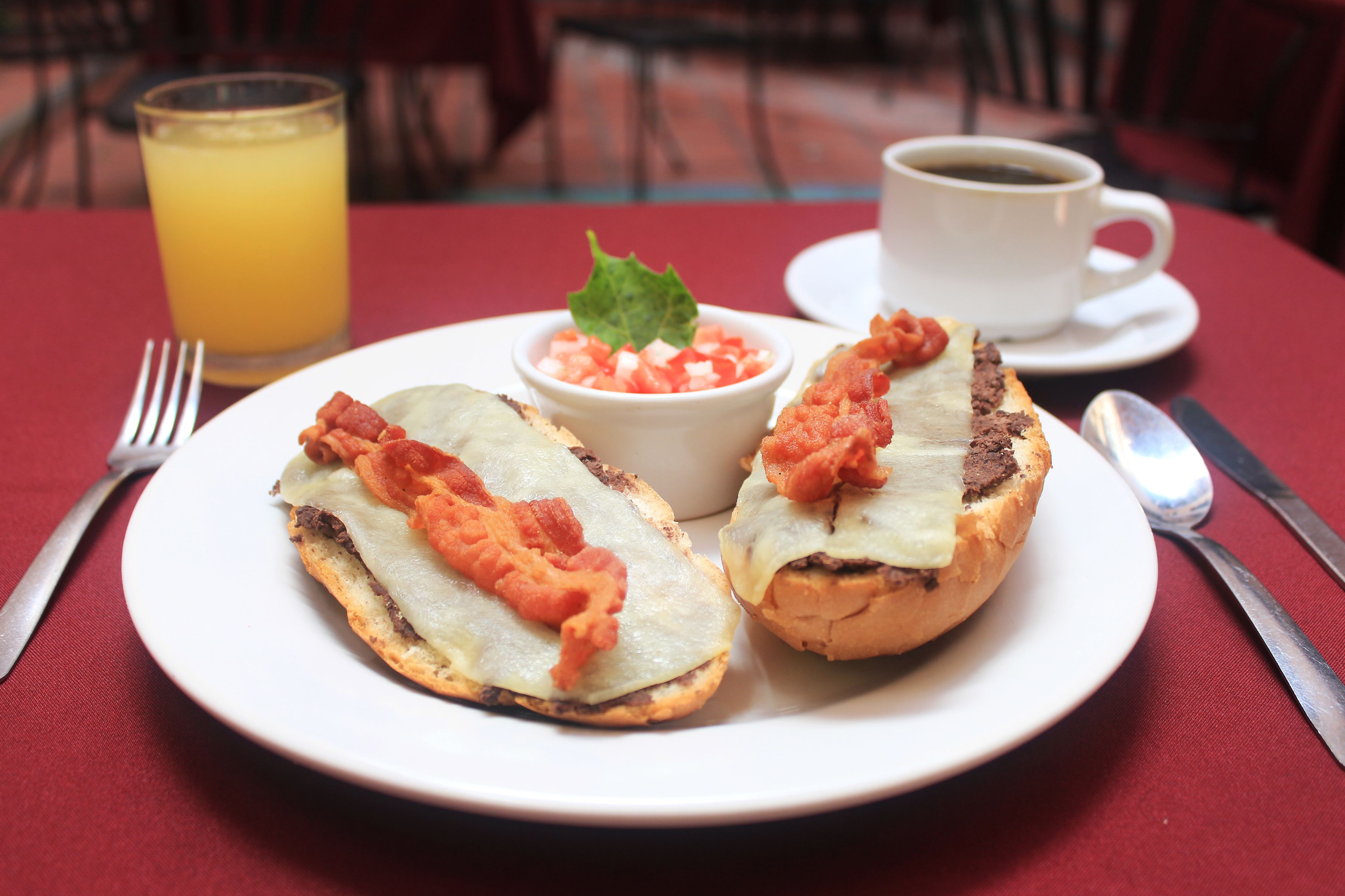 A plate with two sandwiches and a small bowl of diced tomatoes, a glass of orange juice, and a cup of coffee on a red tablecloth.