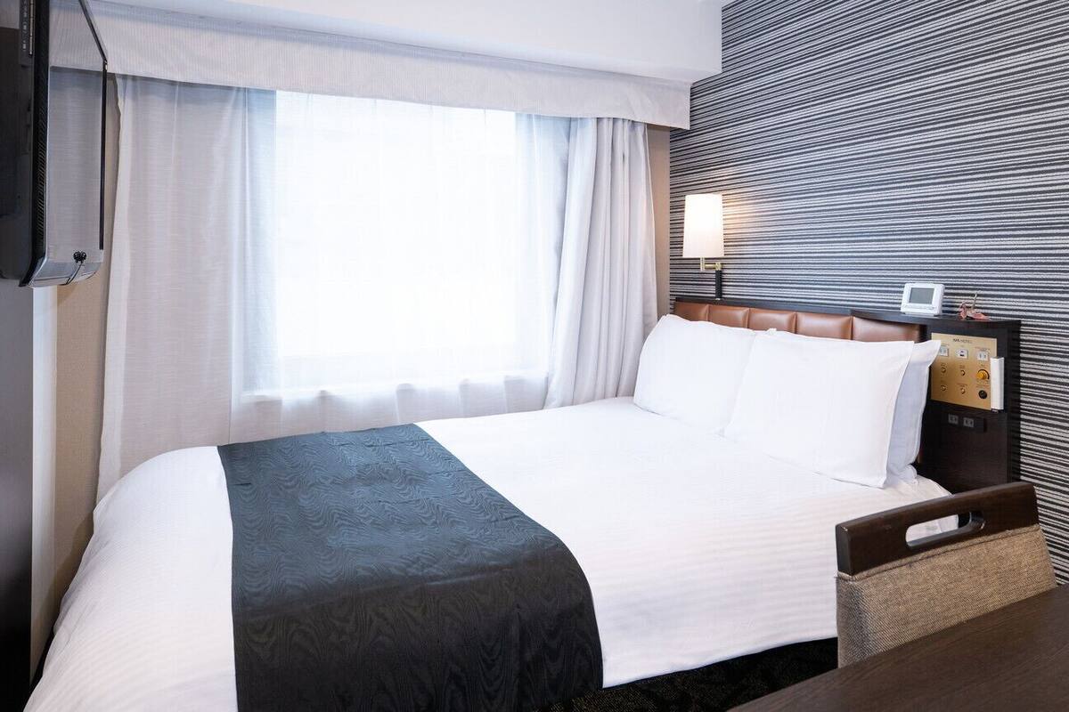 A hotel room with a bed, a bedside table, a lamp, and a wall-mounted control panel.