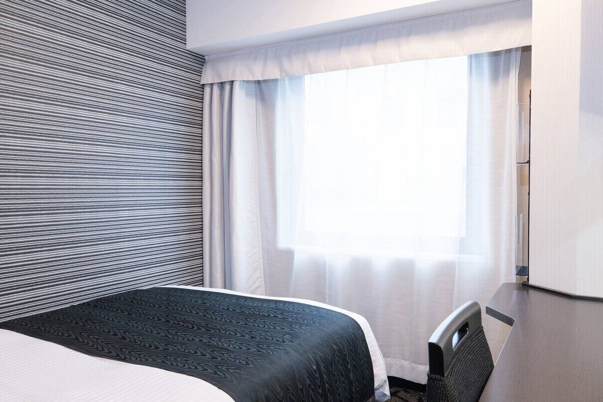 A hotel room with a bed, a chair, a window with curtains, and a striped wall.