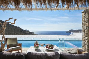 A poolside setting with a view of the sea, a table with a vase, bottles, and a fruit plate. - Myconian Villa Collection - Preferred Hotels & Resorts (Mykonos)