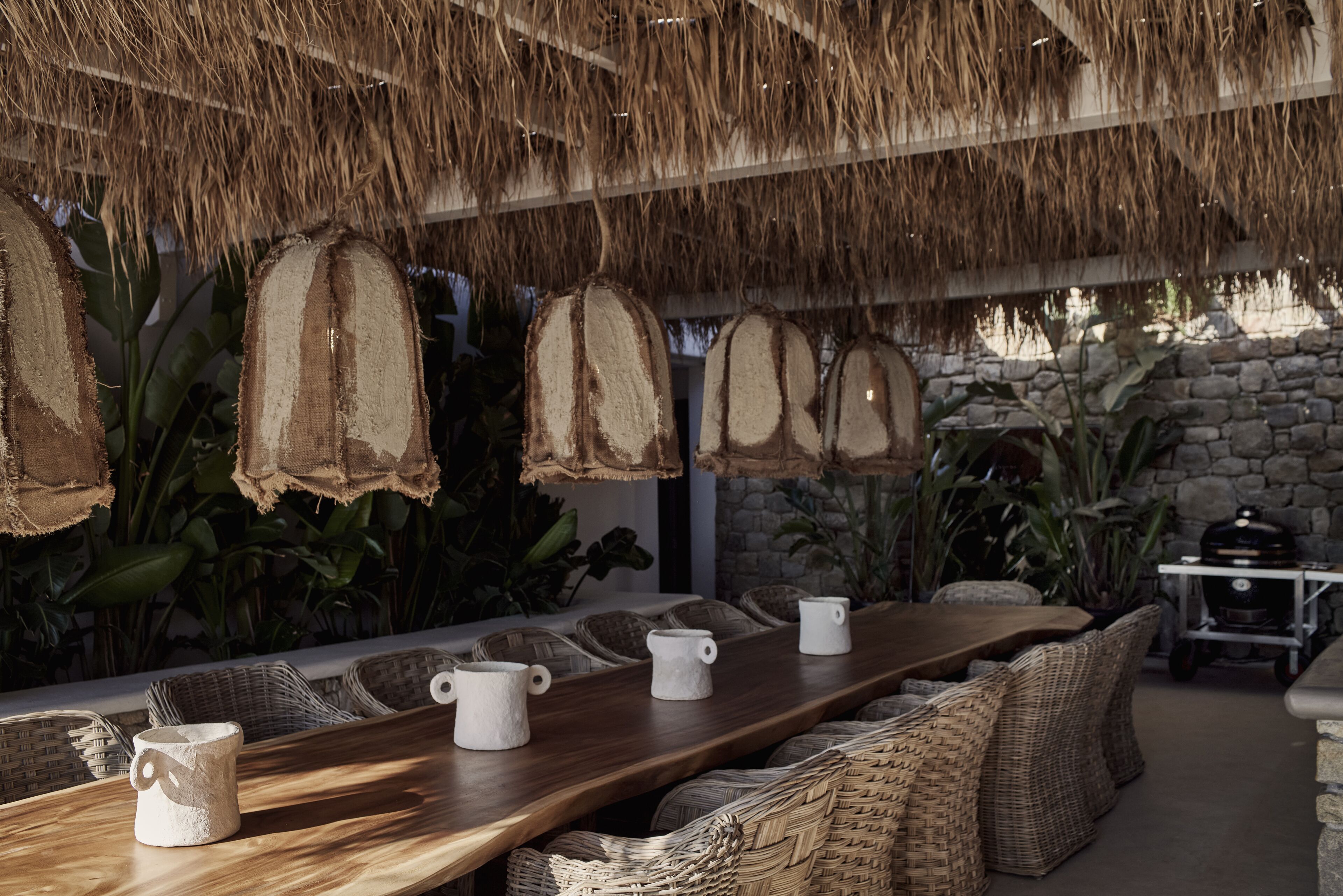 A long wooden table with wicker chairs, surrounded by potted plants and a stone wall.