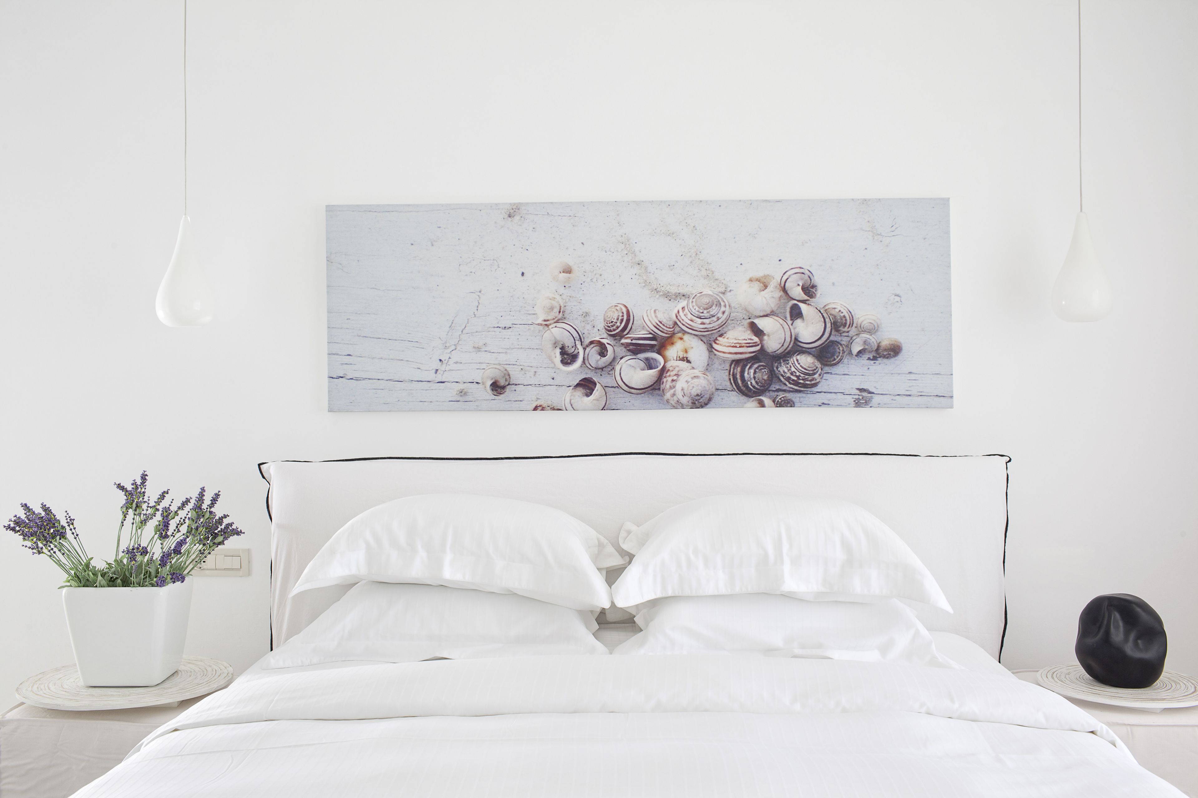 A neatly made bed with white linens, a white potted plant with lavender, and a wall-mounted artwork of seashells.