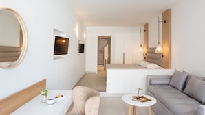 A modern hotel room with a sofa, a small round table, a chair, a bedside table with a glass of water, and a large mirror. - Strogili Hotel - Adults Only (Santorini)