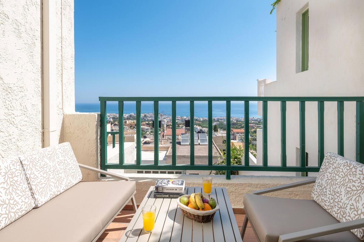Ein Balkon mit Blick auf das Meer, ein Tisch mit einem Buch, ein Glas Orangensaft und ein Obstkorb.