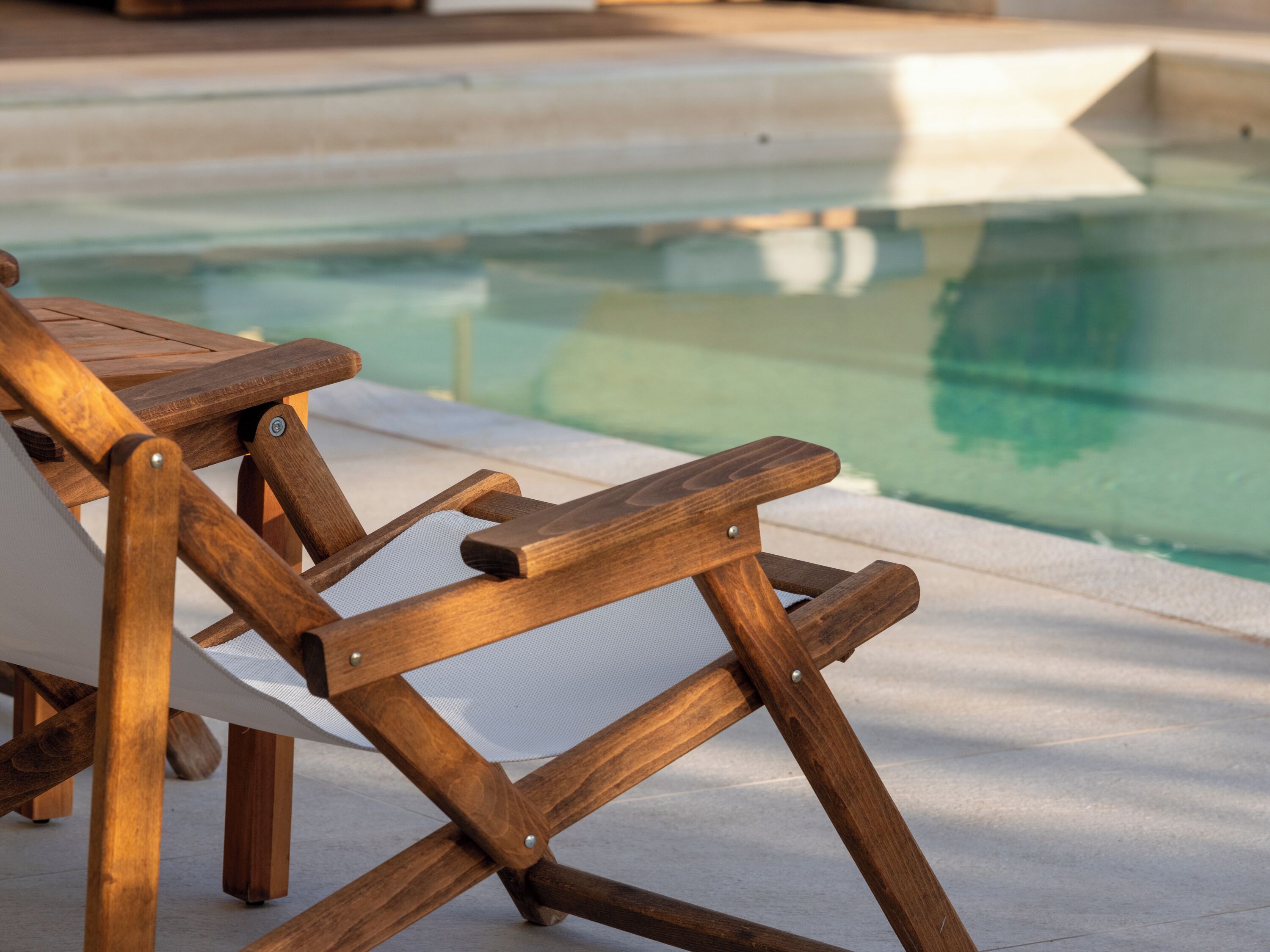A wooden lounge chair with a mesh seat, positioned by a poolside.