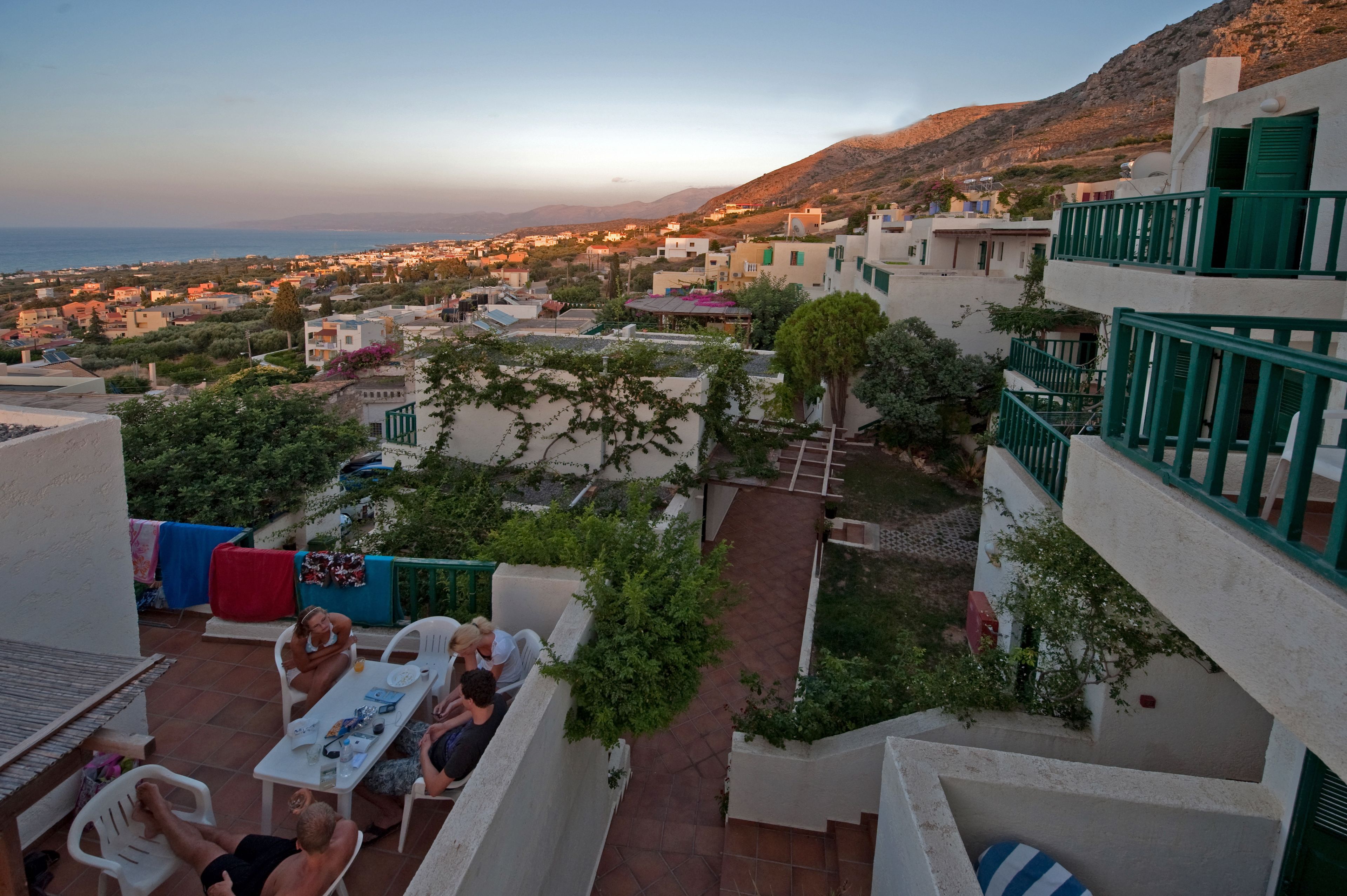 A terrace with outdoor seating, a view of a coastal town, and a hillside in the background.