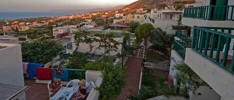 A terrace with outdoor seating, a view of a coastal town, and a hillside in the background.