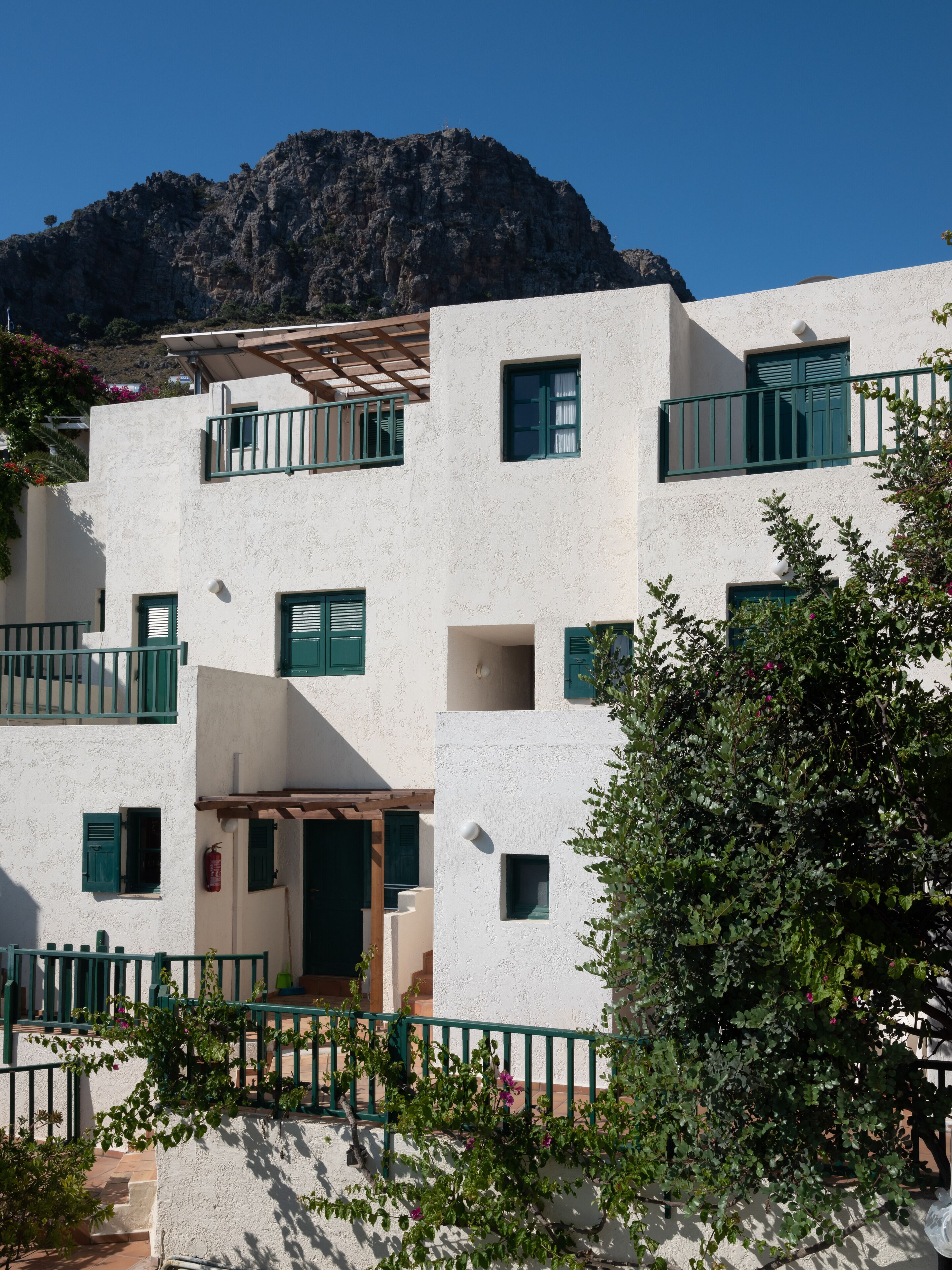 A multi-story building with white walls and green shutters, featuring balconies and a view of a mountain.