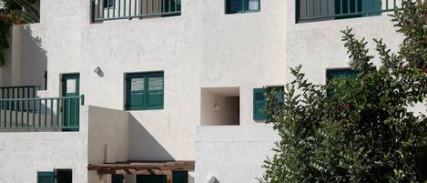 A multi-story building with white walls and green shutters, featuring balconies and a view of a mountain.