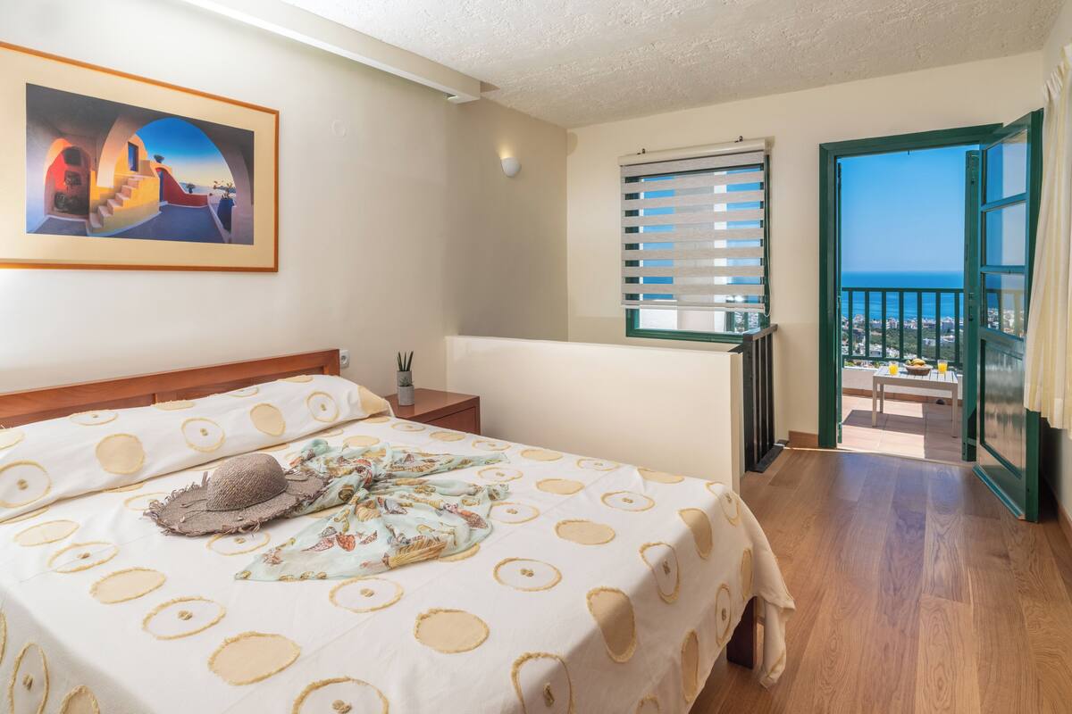 Ein Hotelzimmer mit Bett, einem Gemälde, einem Balkon mit Blick und einem Fenster mit Jalousien.