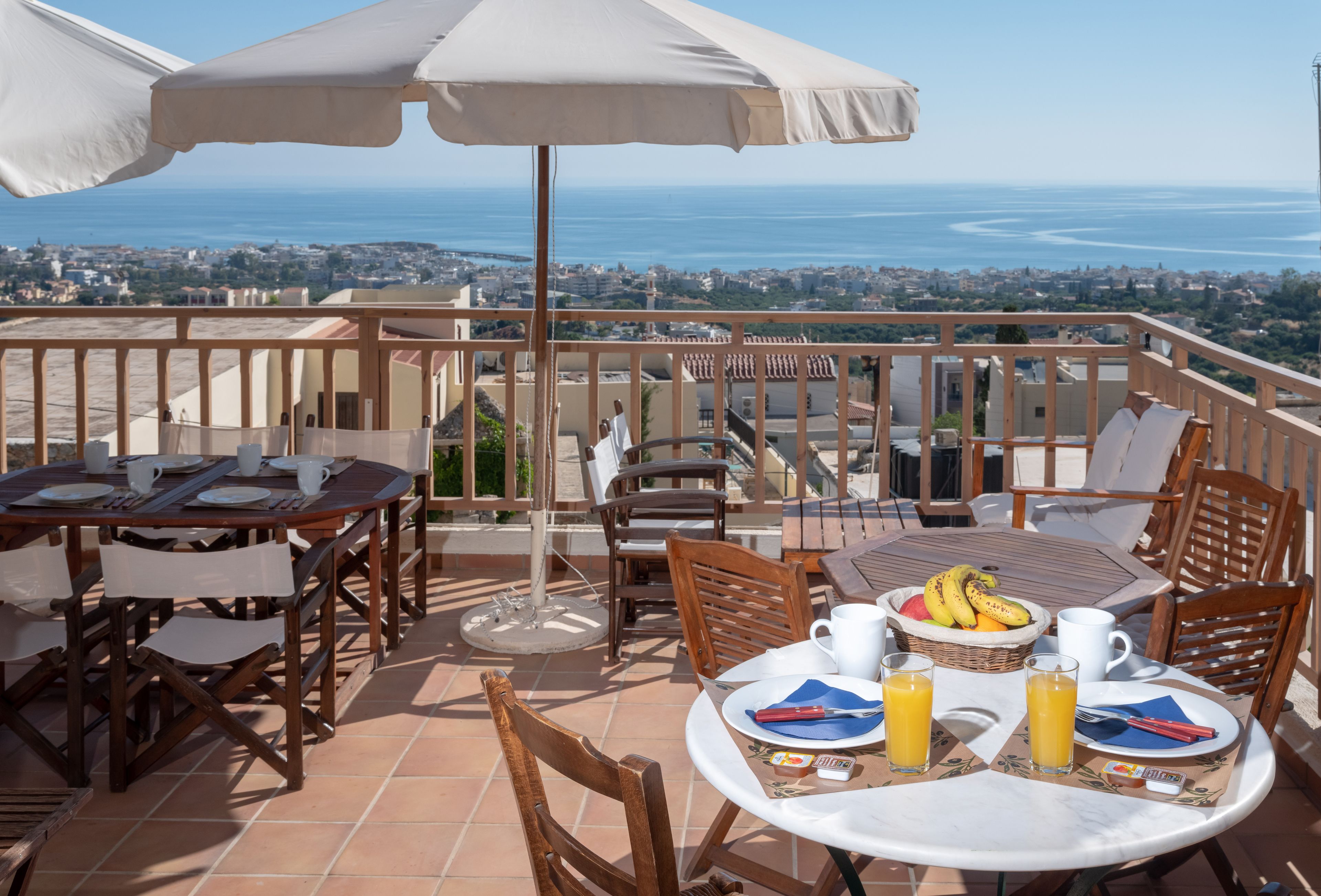 Une terrasse avec des tables et des chaises, une vue sur la mer, et une table dressée avec des assiettes, des tasses et un panier de fruits.