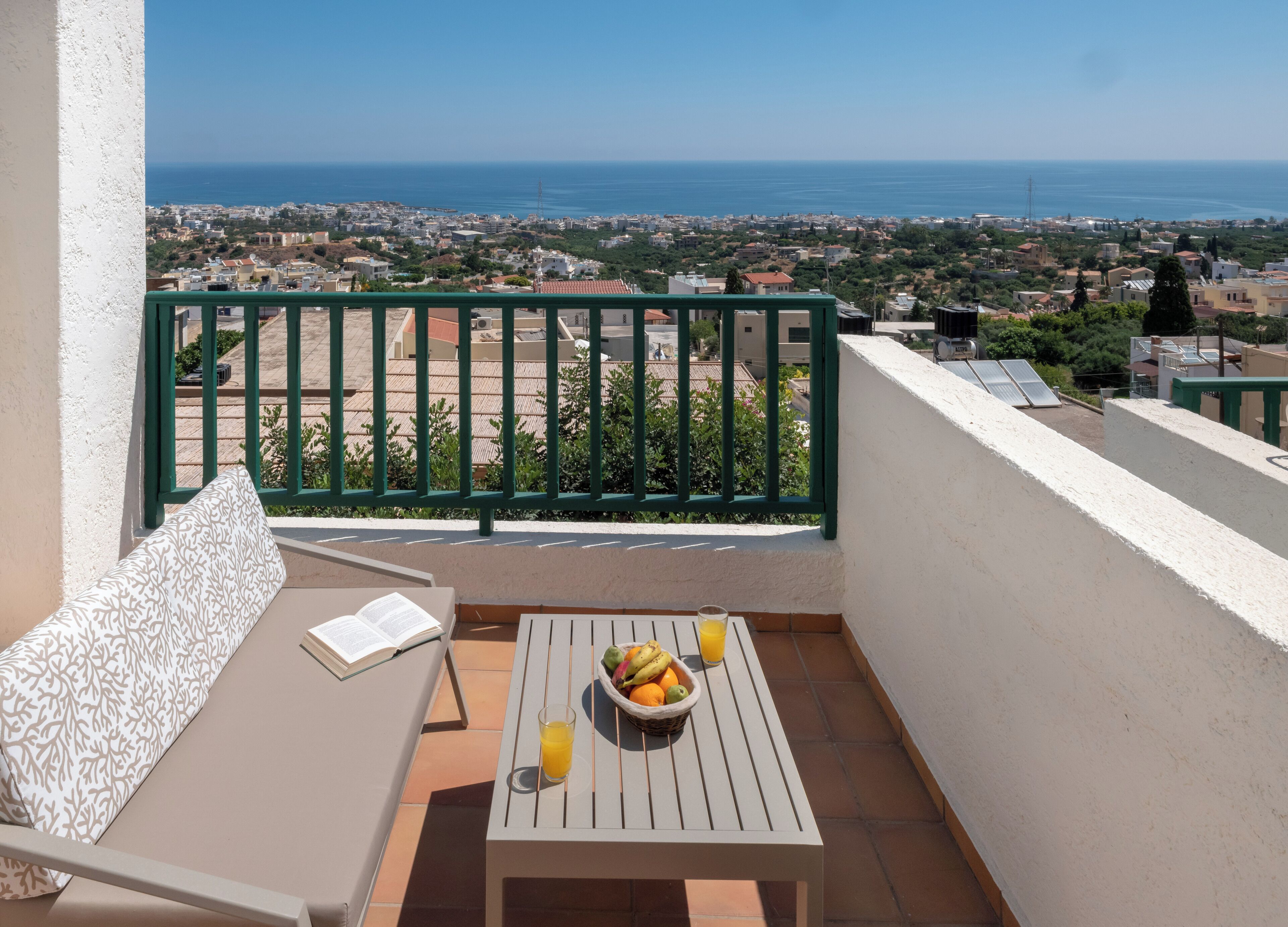 A balcony with a view of the sea, a table with fruit and a drink, and a chair with a book.