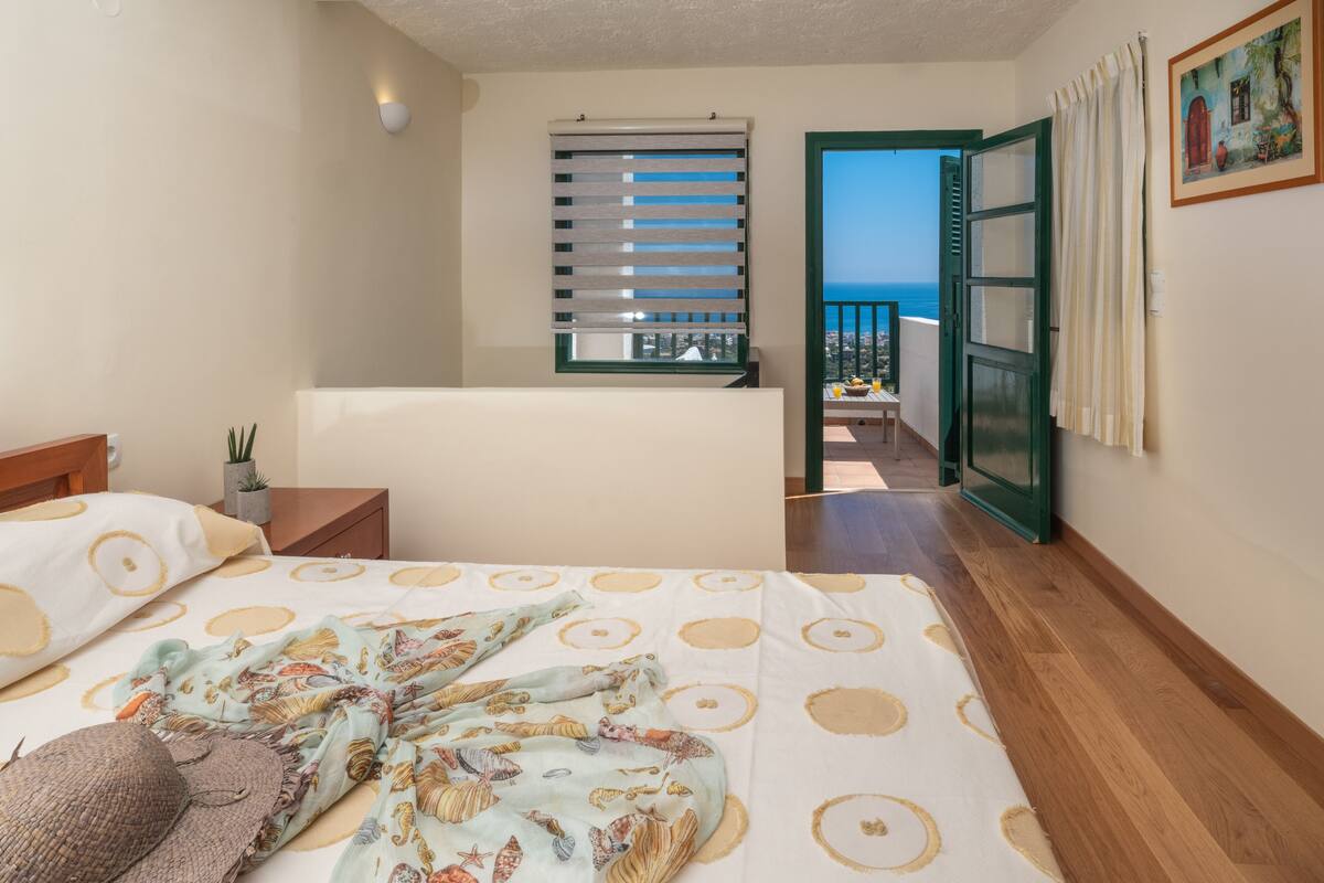 Un dormitorio con cama, ventana con persianas, balcón con vista al mar y puerta que da al exterior.