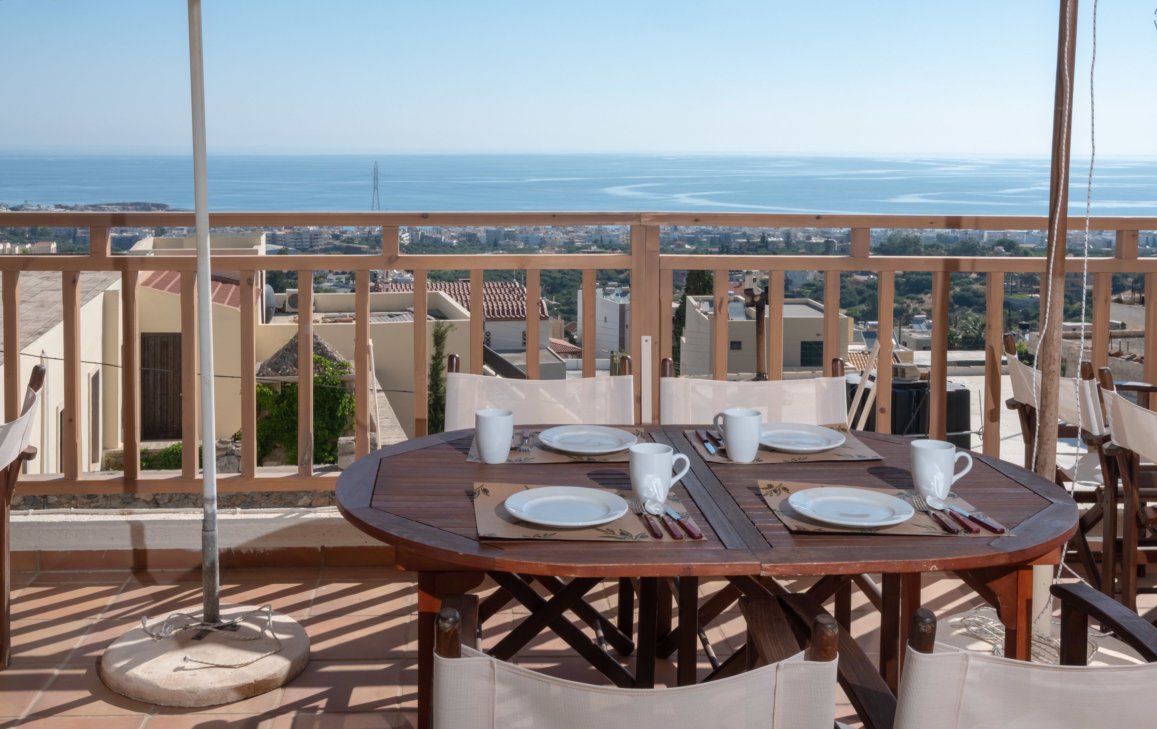 Una mesa de madera con cuatro platos y tazas blancas, colocada en un balcón con vistas al mar y edificios.