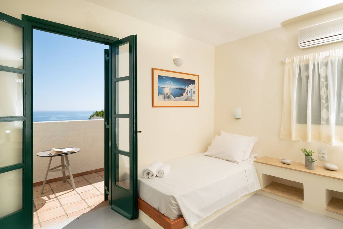 Ein Schlafzimmer mit einem Bett, einem kleinen Tisch, einem Stuhl, einem Bild an der Wand und Meerblick durch eine offene Tür.