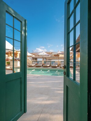 Un espace piscine avec des chaises longues et des parasols, vu à travers des portes ouvertes.