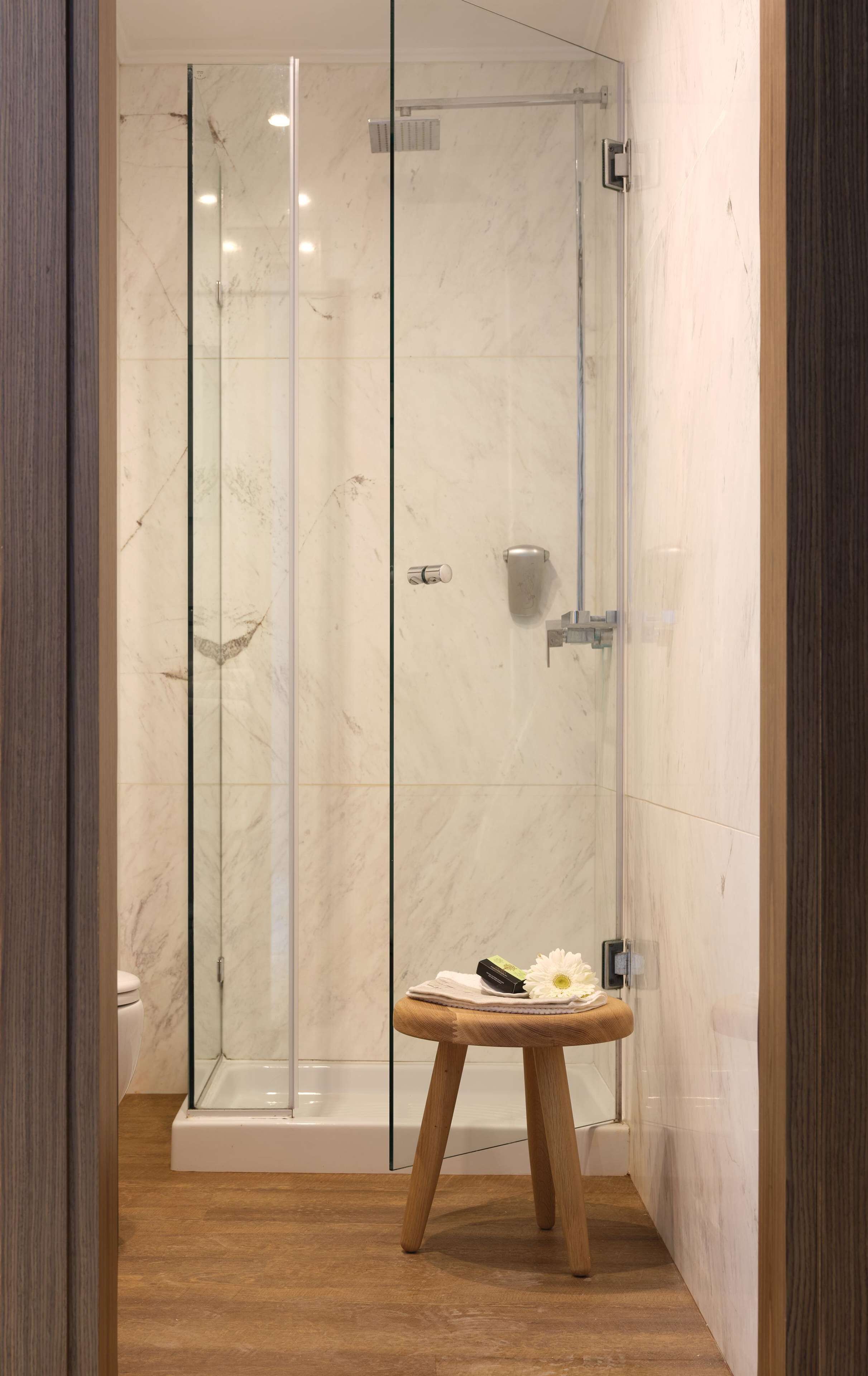 A modern bathroom with a glass-enclosed shower, a wooden stool with toiletries, and a marble-tiled floor.