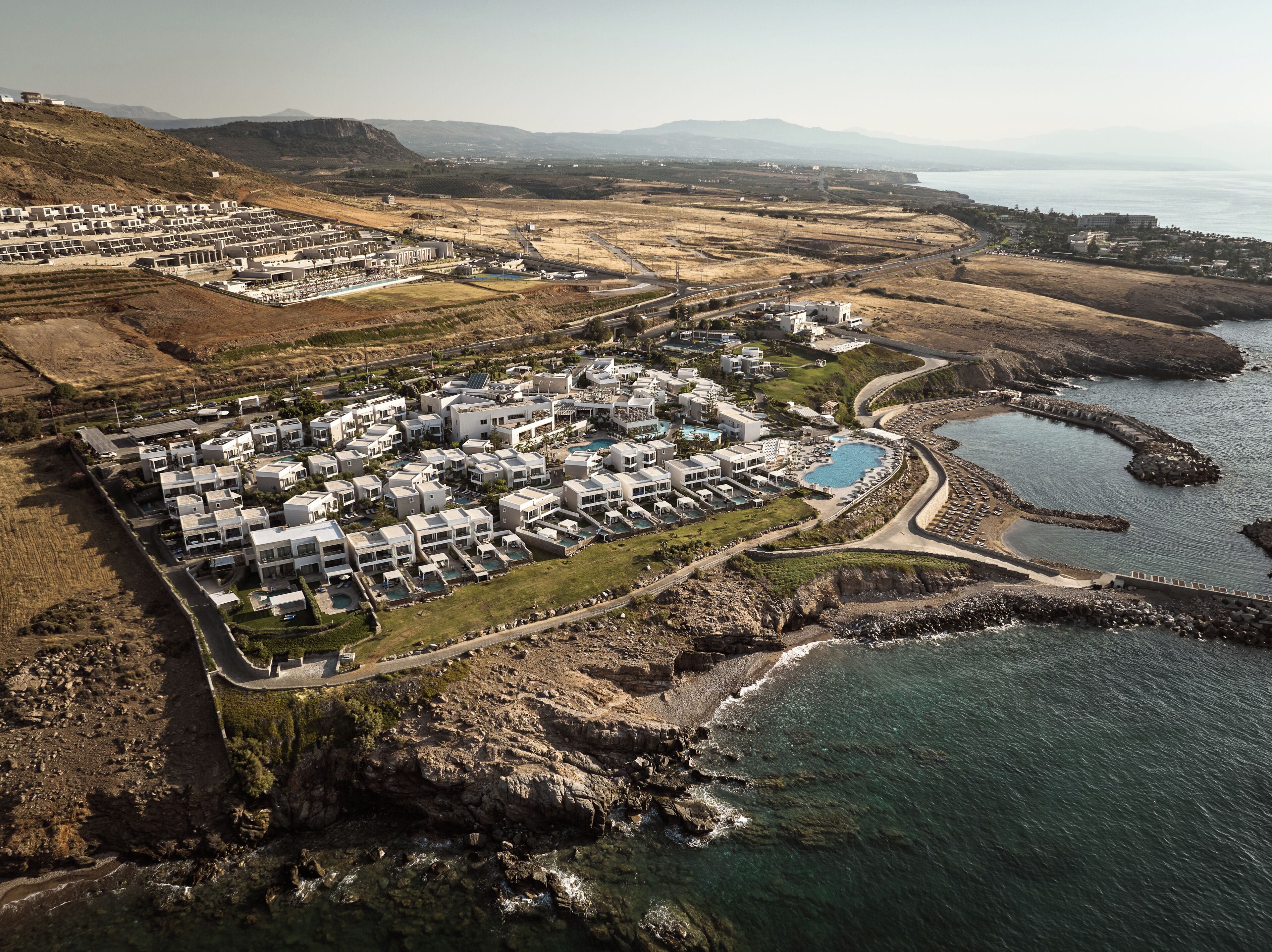 An aerial view of a coastal resort with multiple buildings, a swimming pool, and a rocky shoreline.