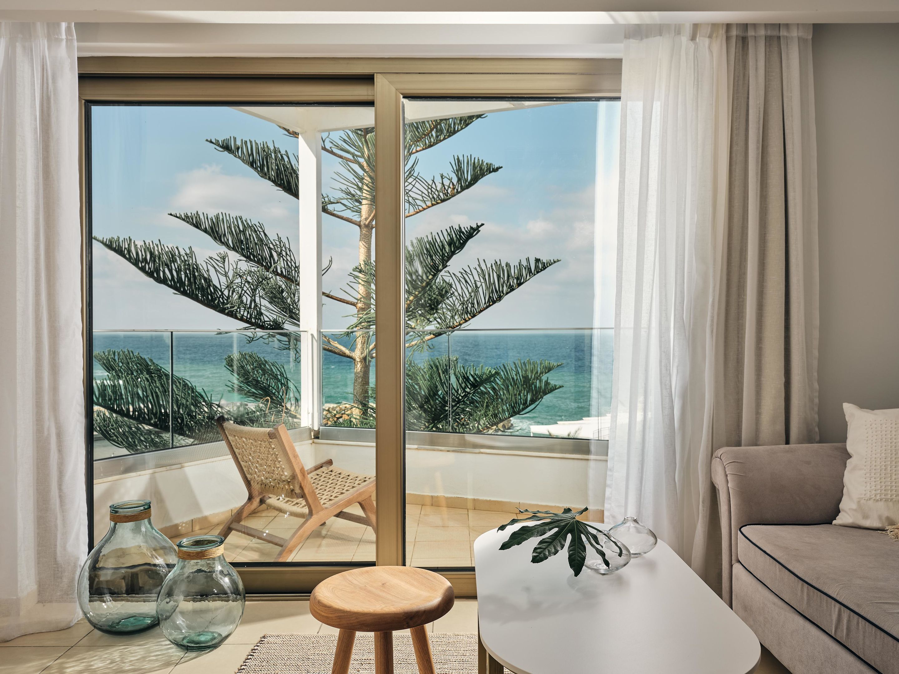 Un balcon avec vue sur l’océan et les palmiers, une chaise en bois, une petite table avec une plante et un canapé.