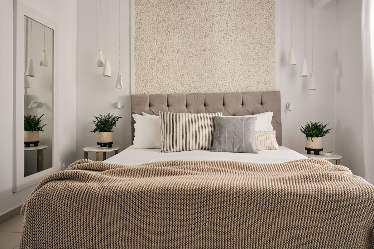 A modern bedroom with a tufted headboard, a large bed with multiple pillows, bedside tables with plants, and pendant lighting.