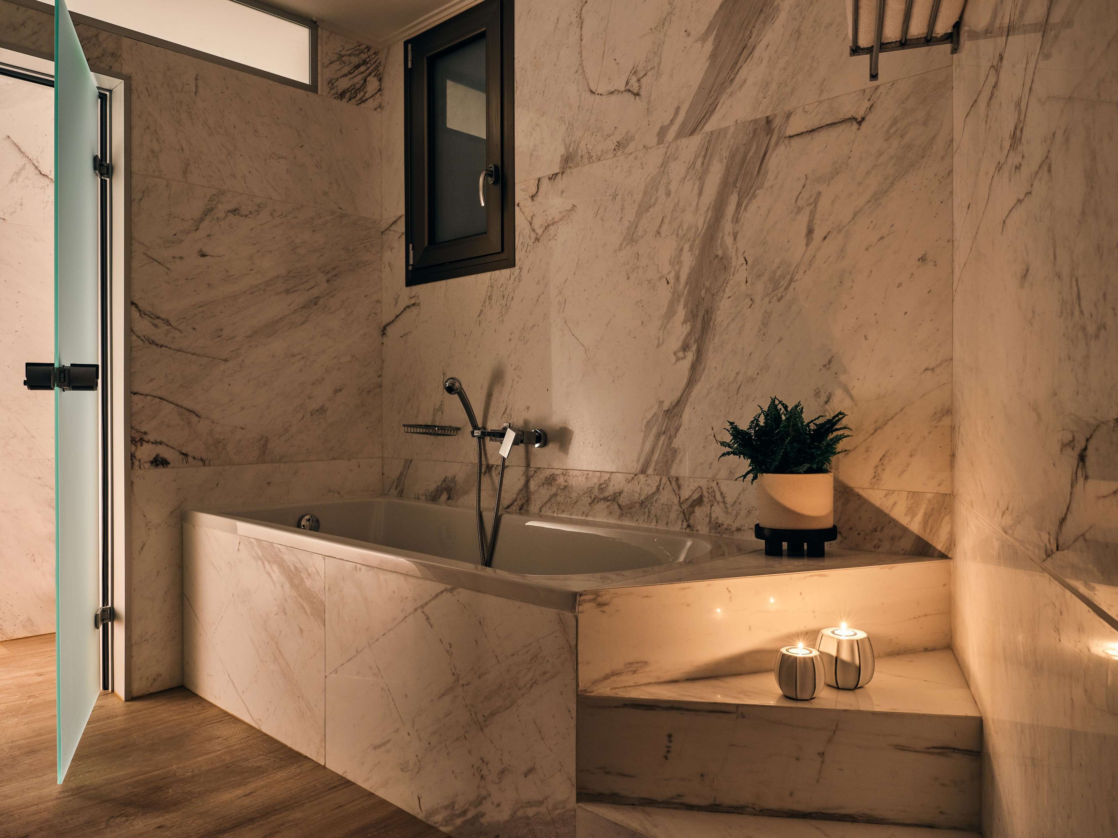Ein modernes Badezimmer mit Marmorwand, einer Badewanne, einer Topfpflanze und Kerzen.
