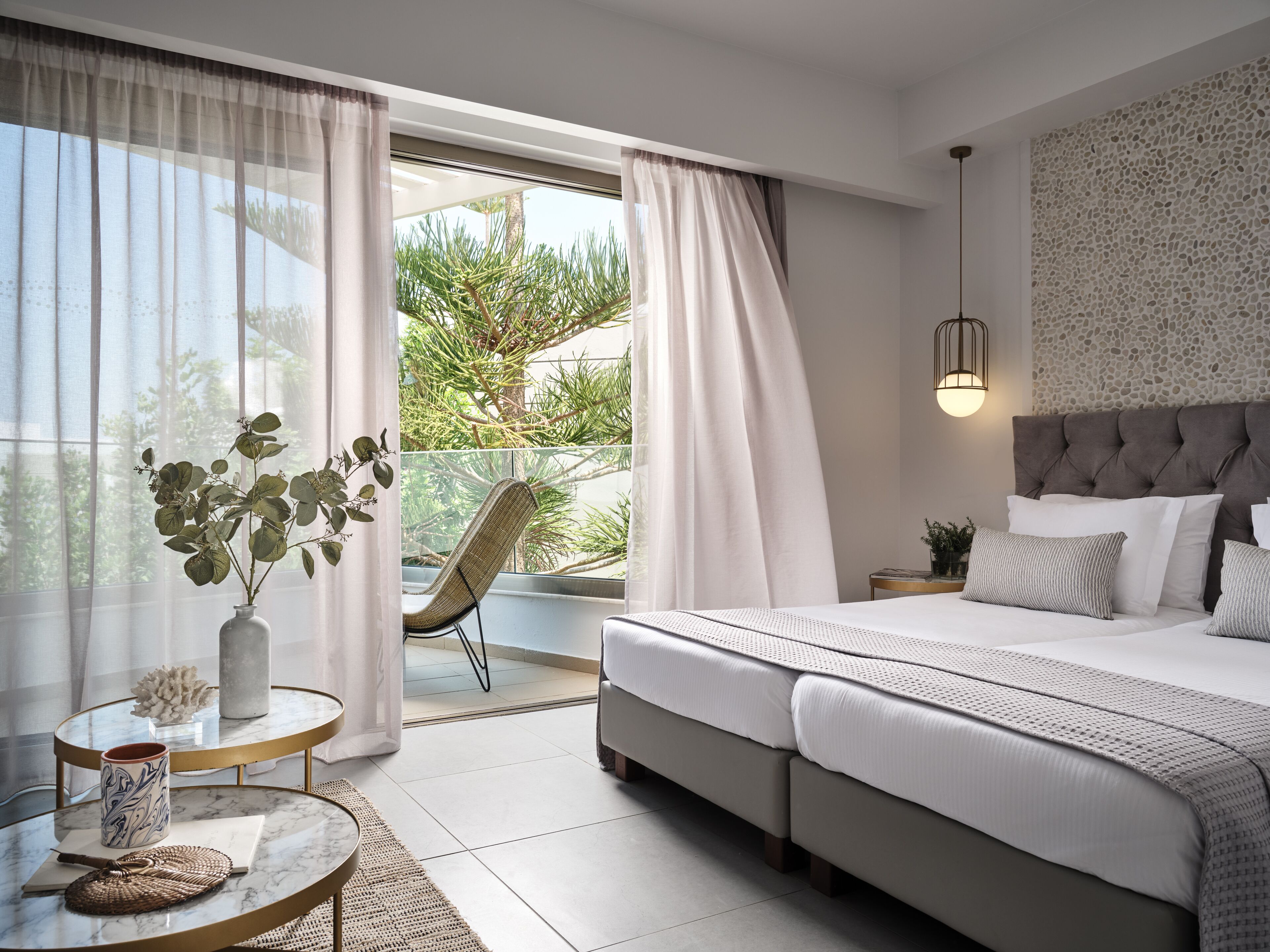 Un dormitorio moderno con una cama grande, una zona de estar con una silla, una mesita con un florero y una taza, y vistas a zonas verdes al exterior.