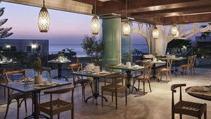 An outdoor dining area with tables and chairs, hanging lights, and a view of the ocean. - The Royal Blue (Mylopotamos)