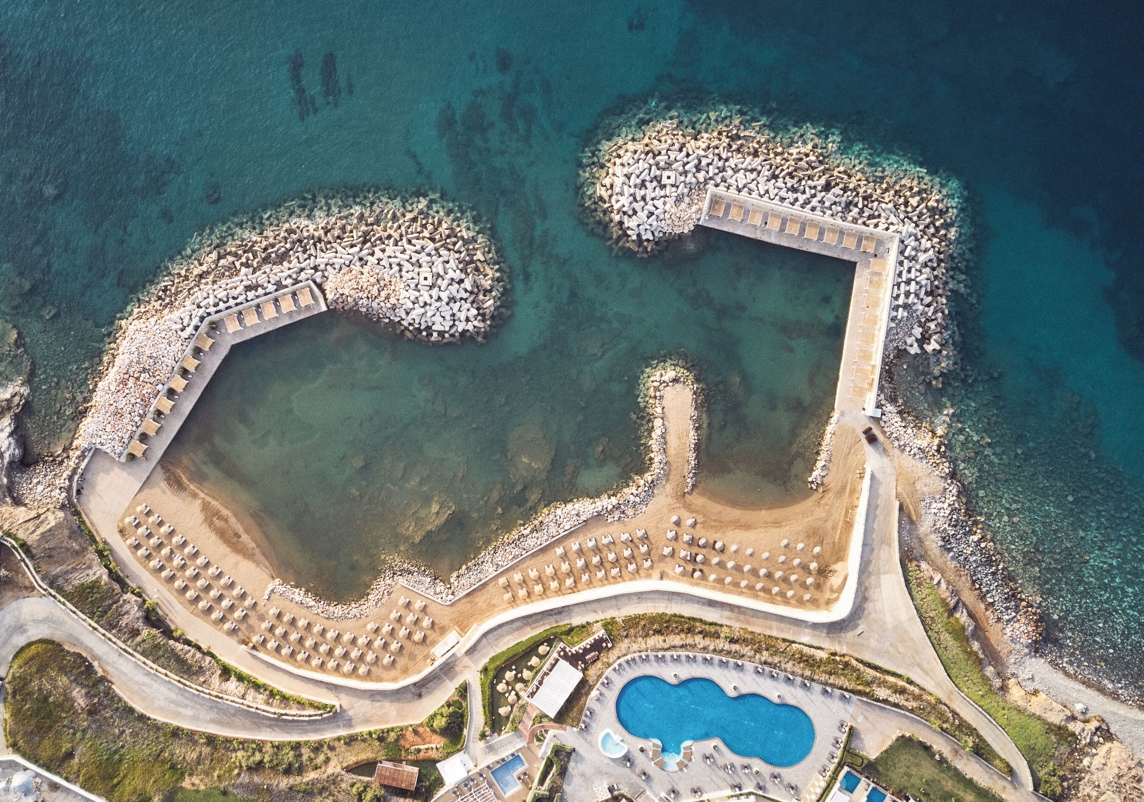 Luftaufnahme eines Küstenresorts mit Pool, Strand und felsiger Küstenlinie.