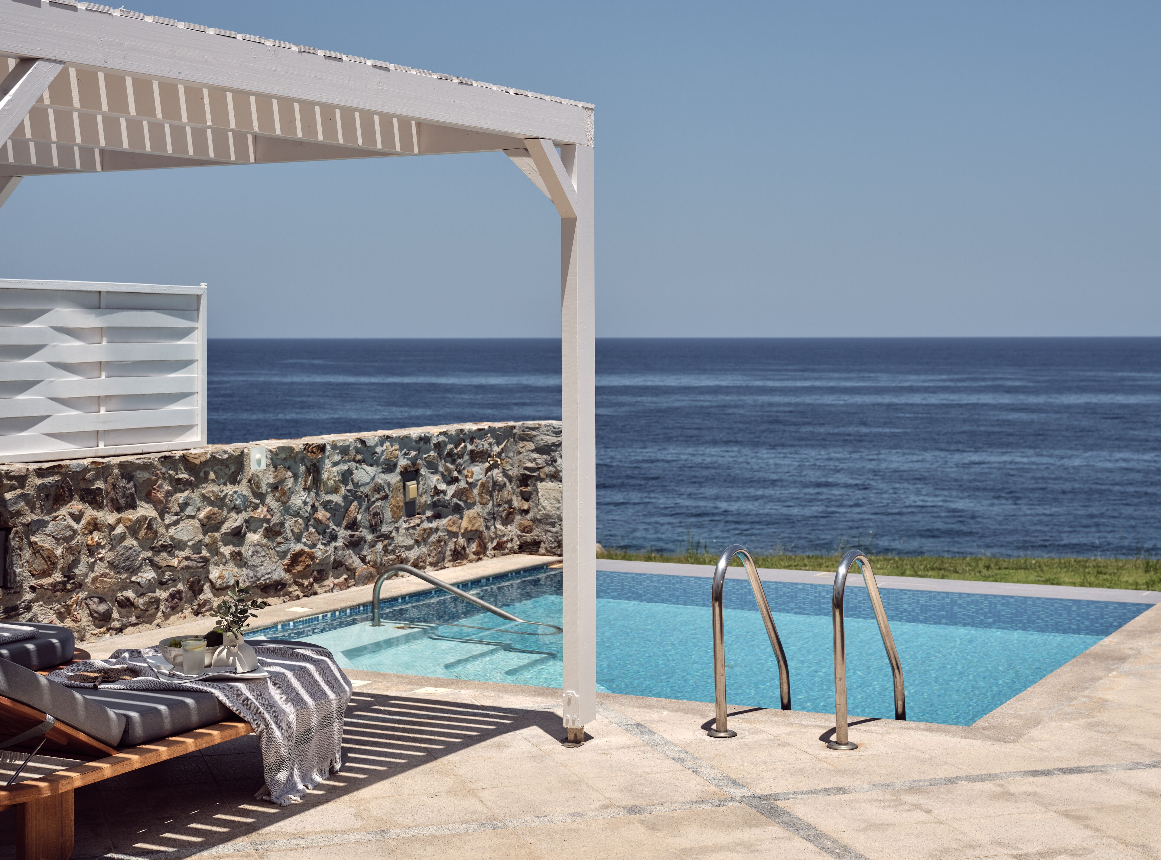 A pool area with a stone wall, lounge chairs, and a view of the ocean.