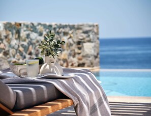 A poolside lounge with a stone wall, a potted plant, and a glass of water with a lemon slice. - The Royal Blue (Mylopotamos)