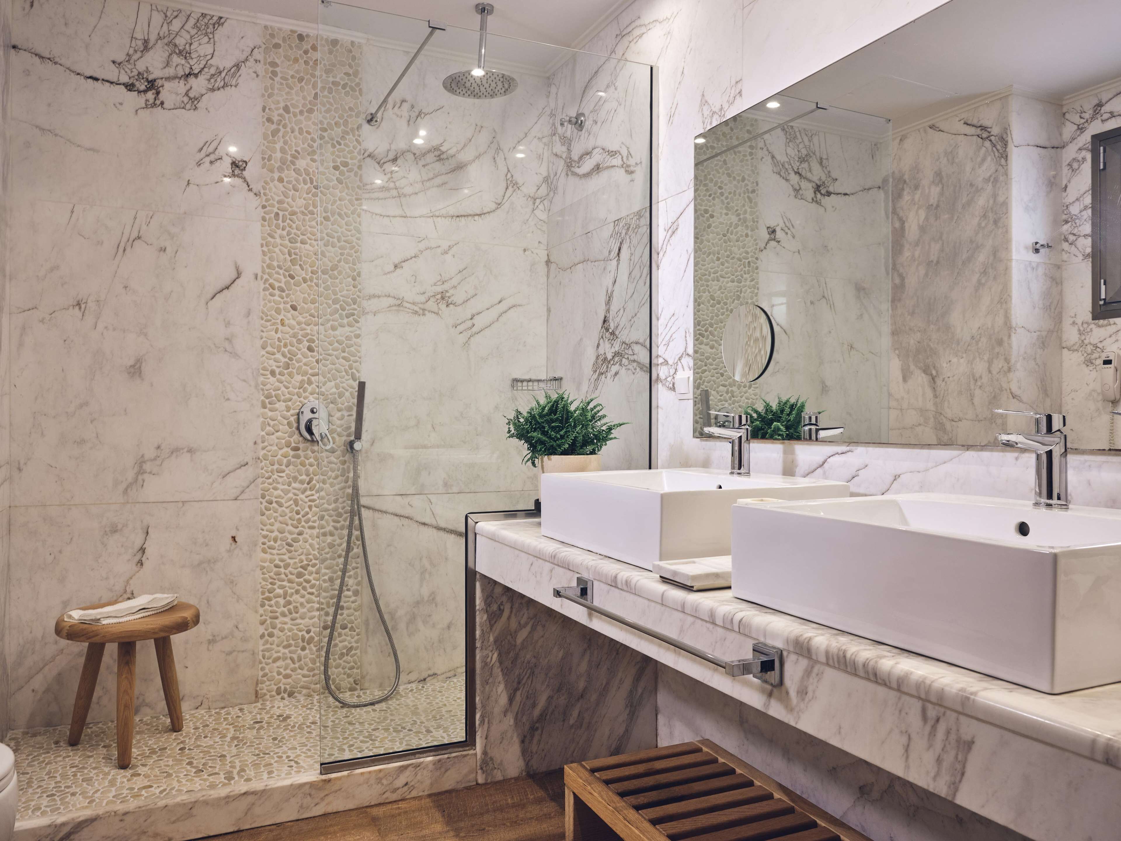 A modern bathroom with a glass-enclosed shower, two sinks, a large mirror, and a wooden stool.