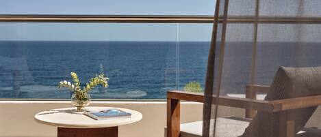 A balcony with a view of the ocean, a round table with a vase and a book, and a chair with a cushion.