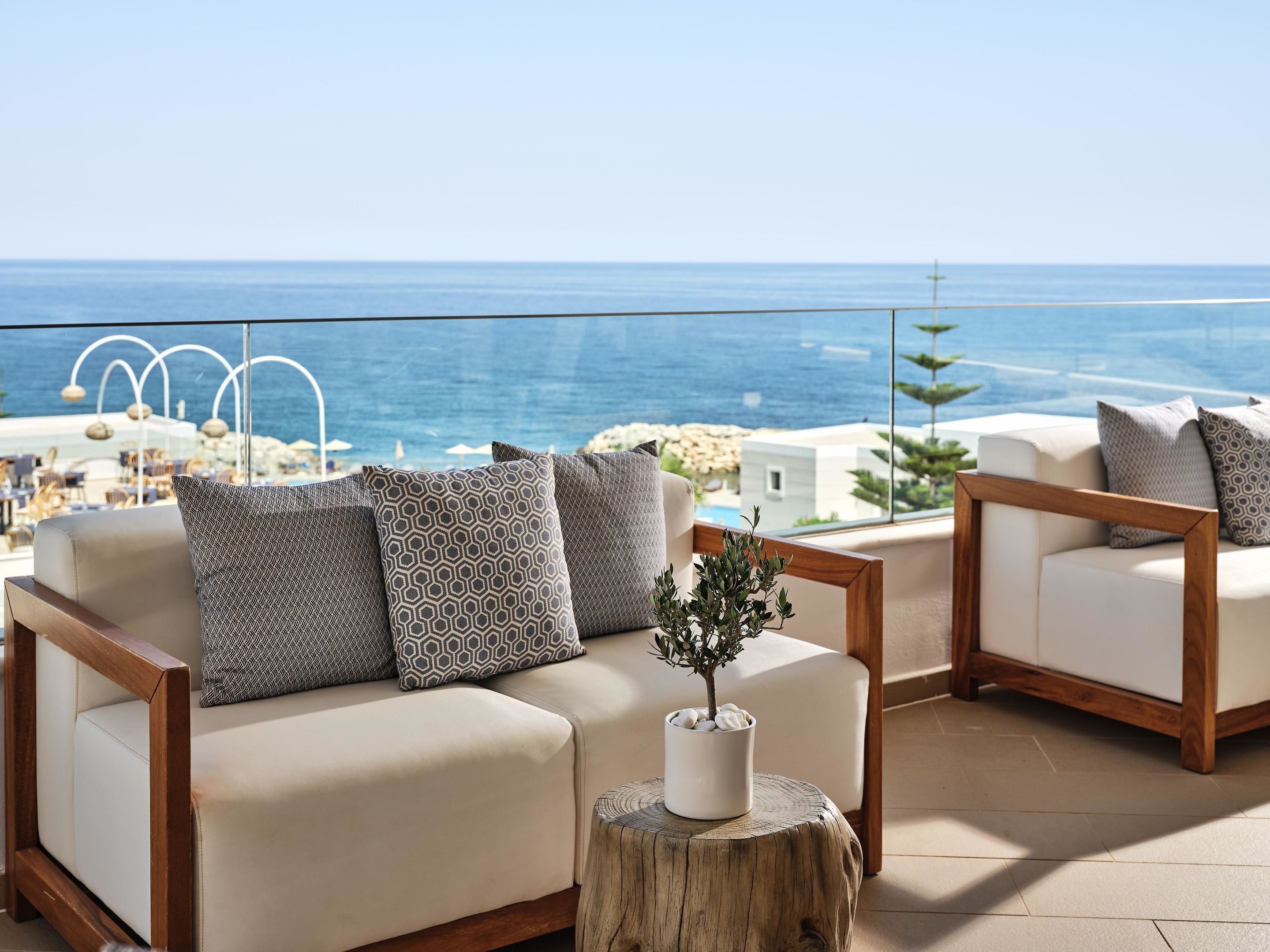 Een balkon met een bank en fauteuils, een houten krukje met een plant, en uitzicht op zee en gebouwen.