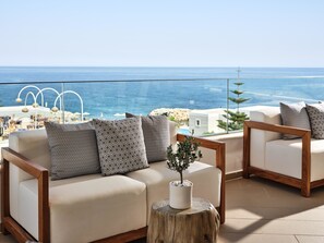 A balcony with a sofa and armchairs, a wooden stool with a plant, and a view of the sea and buildings. - The Royal Blue (Mylopotamos)