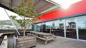 Un espace extérieur aménagé avec des bancs en bois et un arbre, de grandes fenêtres et une façade rouge.