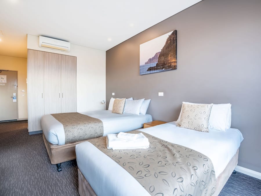 A hotel room with two beds, a wardrobe, an air conditioning unit, and a painting on the wall.