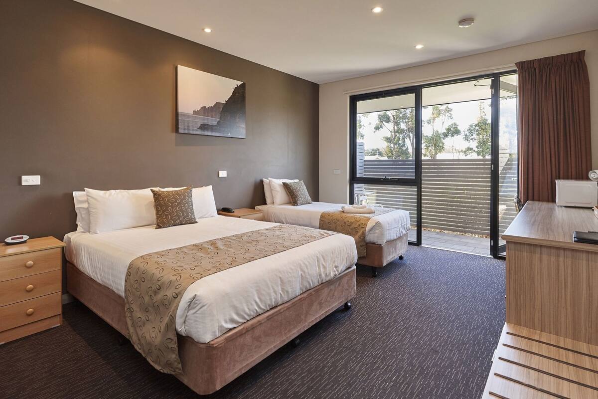A hotel room with two beds, a desk, a chair, a window with a view, and a painting on the wall.