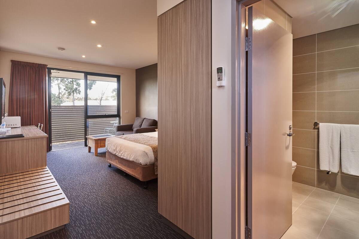 A modern hotel room with a bed, a desk, a chair, a small table, and a shower area.