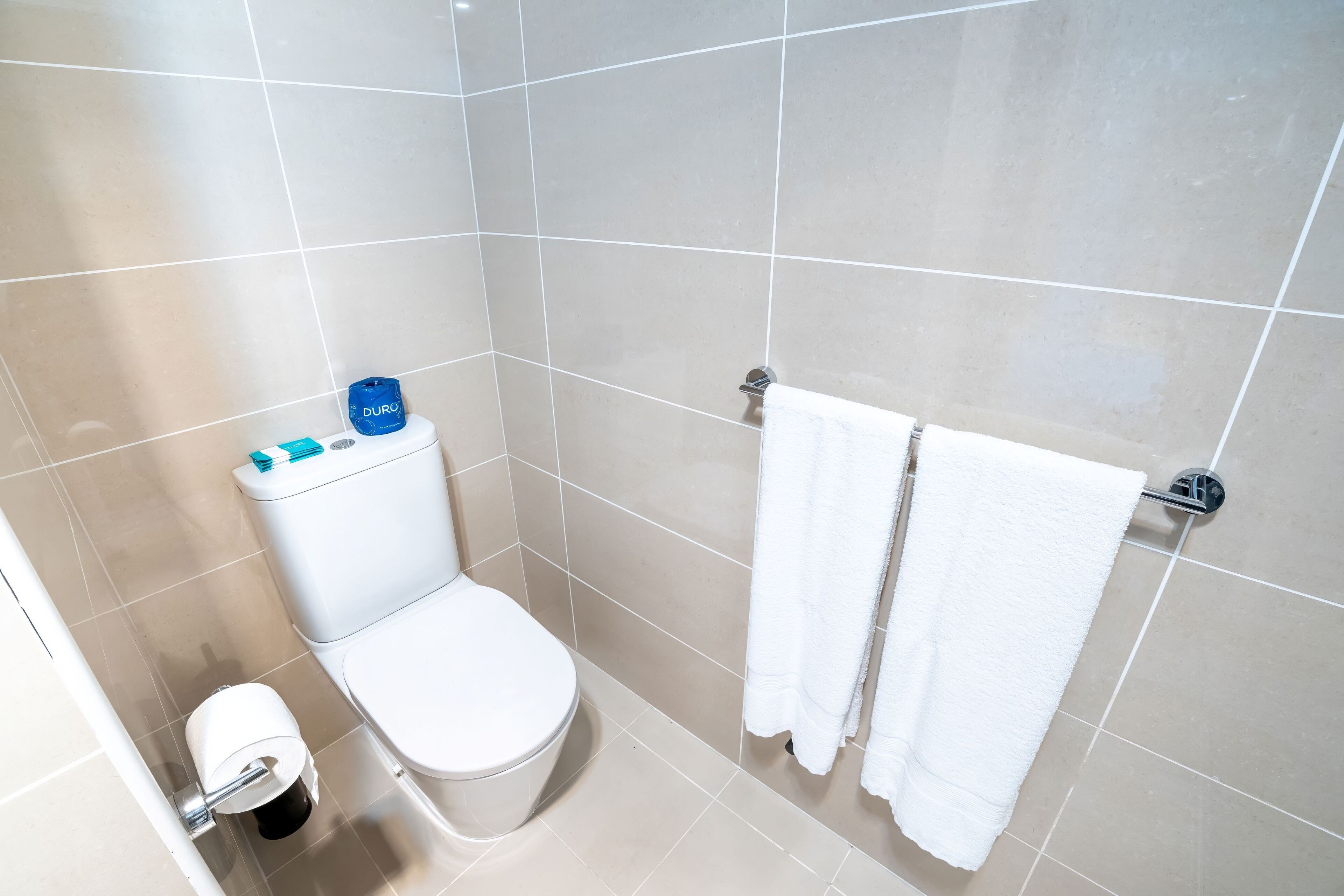 A bathroom with a toilet, a roll of toilet paper, a blue water bottle, and a towel rack with two white towels.