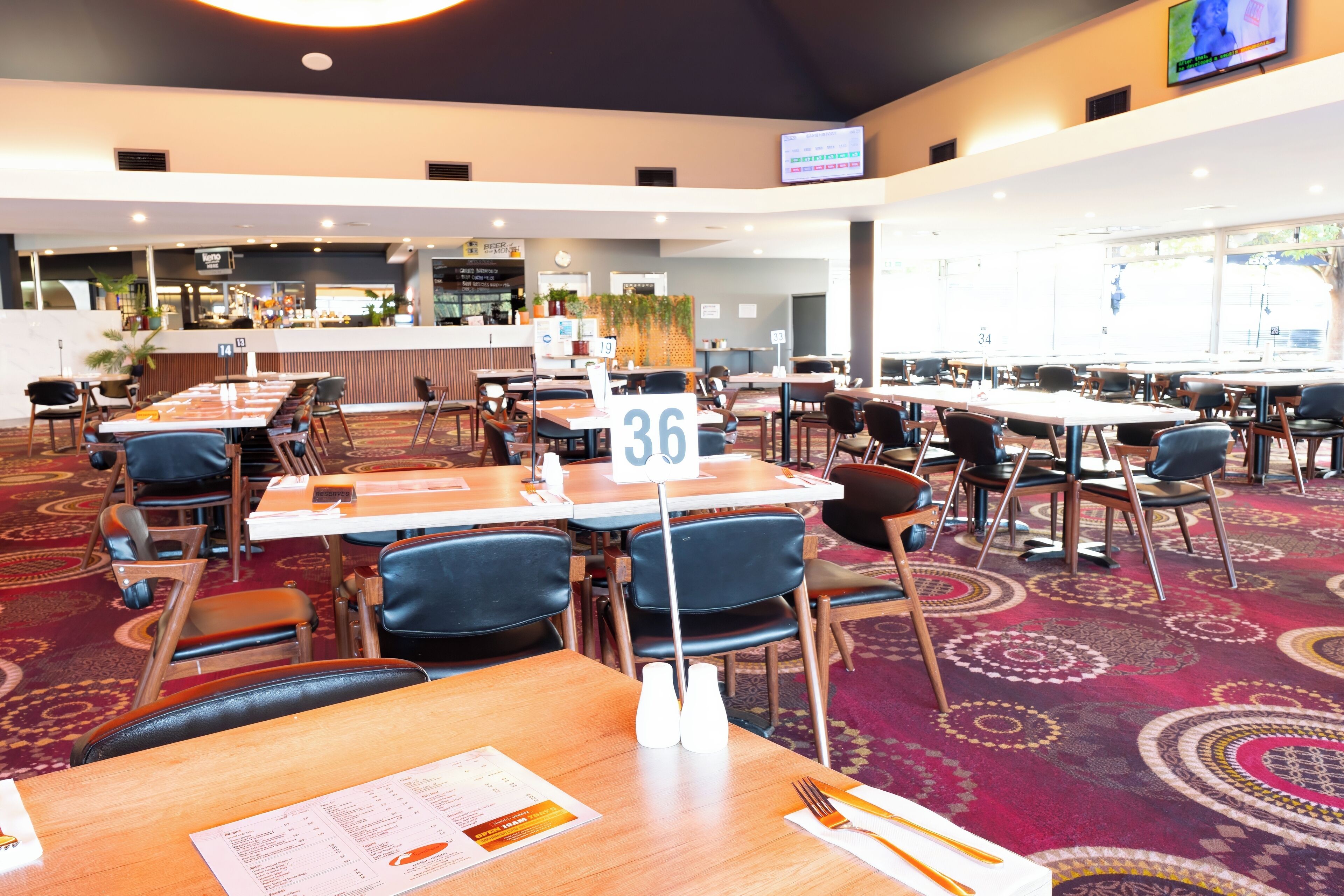 A hotel dining area with multiple tables and chairs, a counter with a menu, and a television screen mounted on the wall.