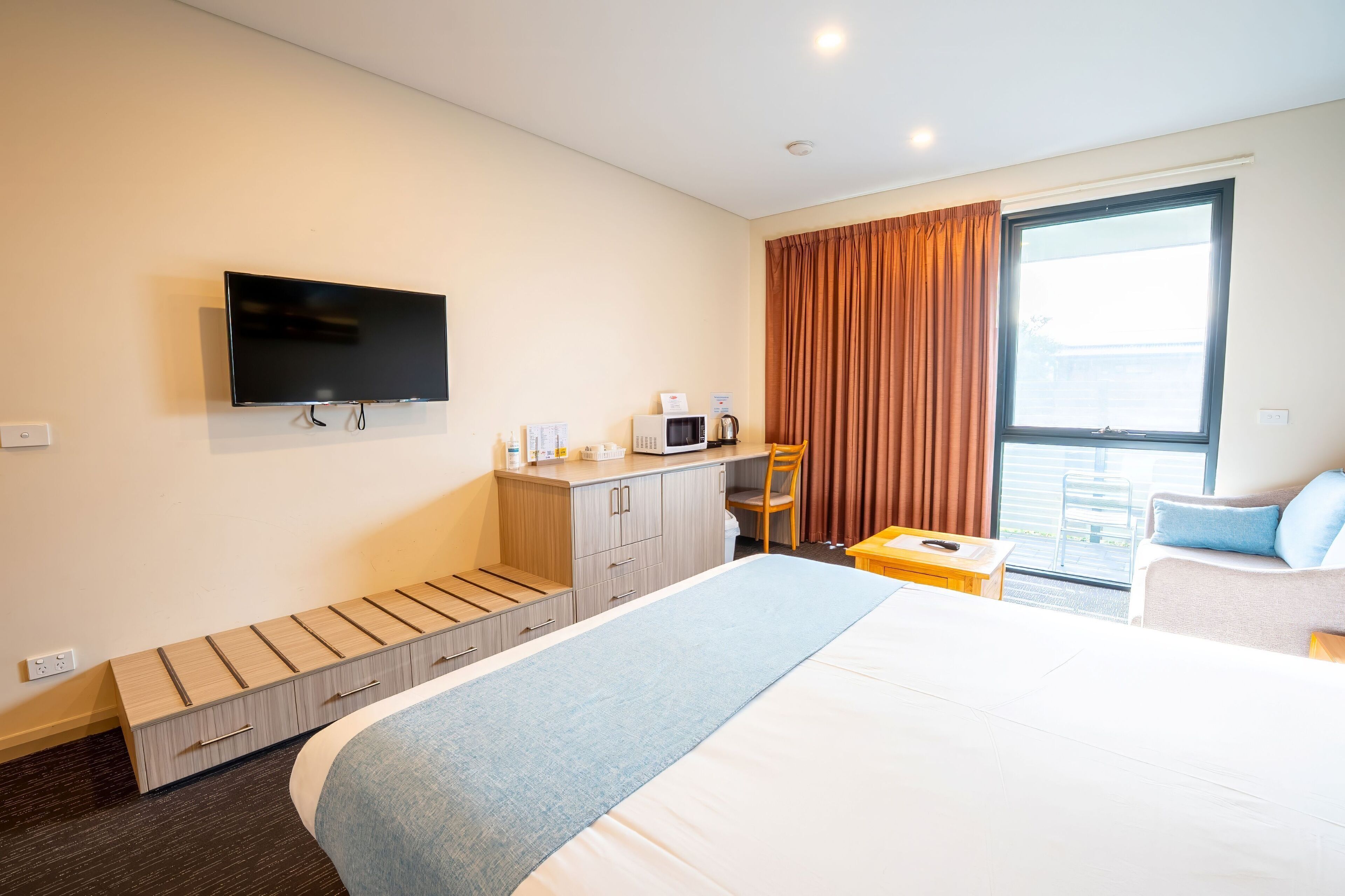 Ein Hotelzimmer mit Bett, einem an der Wand befestigten Fernseher, einem Schreibtisch mit eingebauter Mikrowelle und Blick ins Freie durch ein großes Fenster.