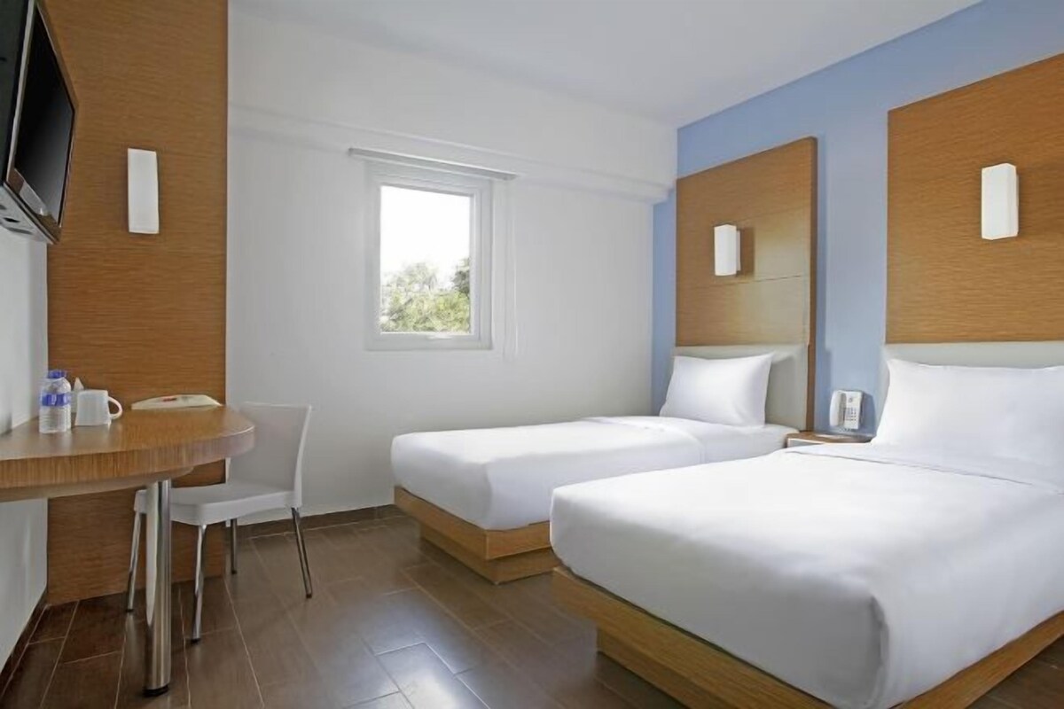 Une chambre d’hôtel avec deux lits simples, un bureau en bois avec une chaise, une télévision et une fenêtre.