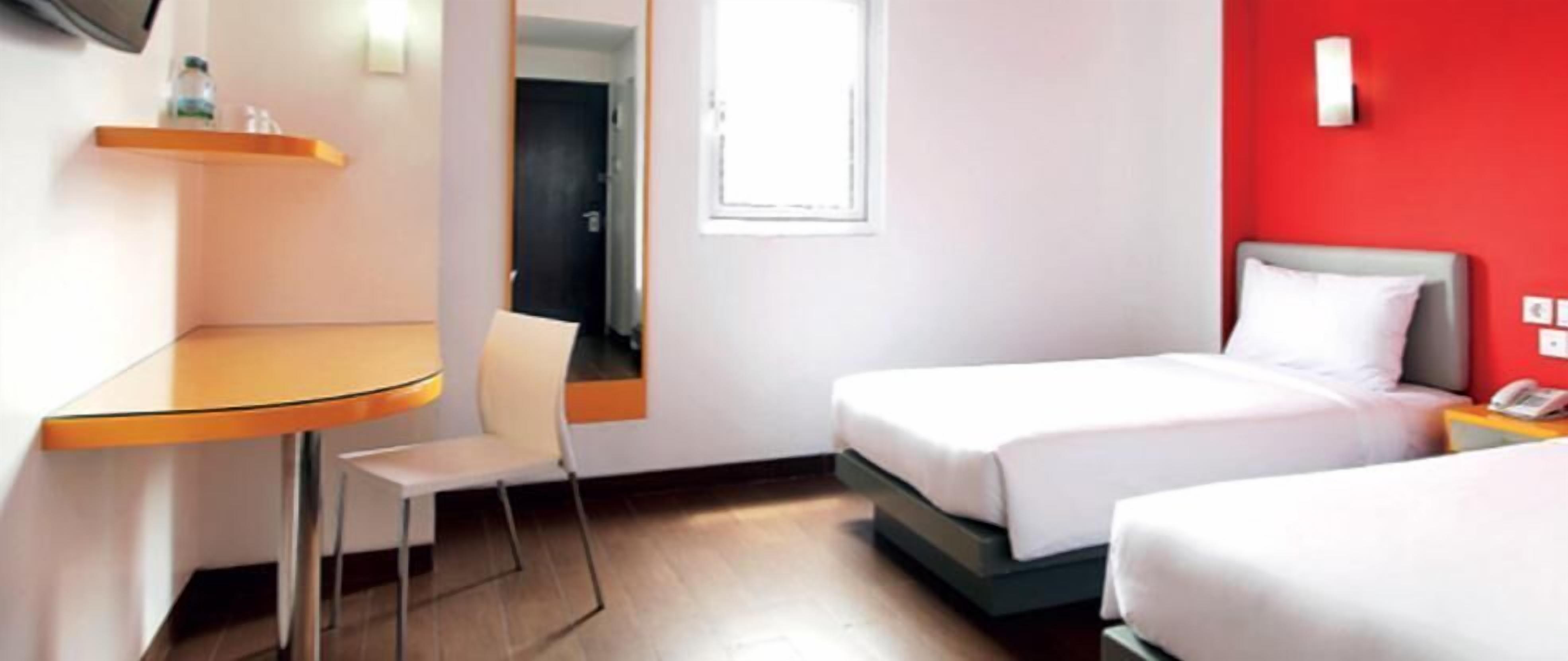 Hotellihuone, jossa on kaksi sänkyä, pieni pöytä, tuoli ja punainen tehosteseinä.