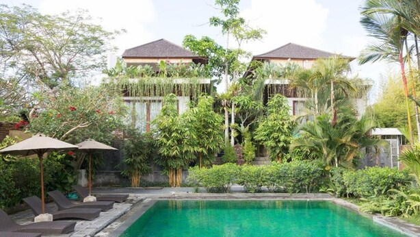 A pool area with lounge chairs and umbrellas, surrounded by lush greenery and a multi-story building with a tiled roof.