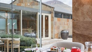 A modern outdoor dining area with a view of a mountain, stone walls, and a glass door leading to a patio. - Islington Hotel (South Hobart)