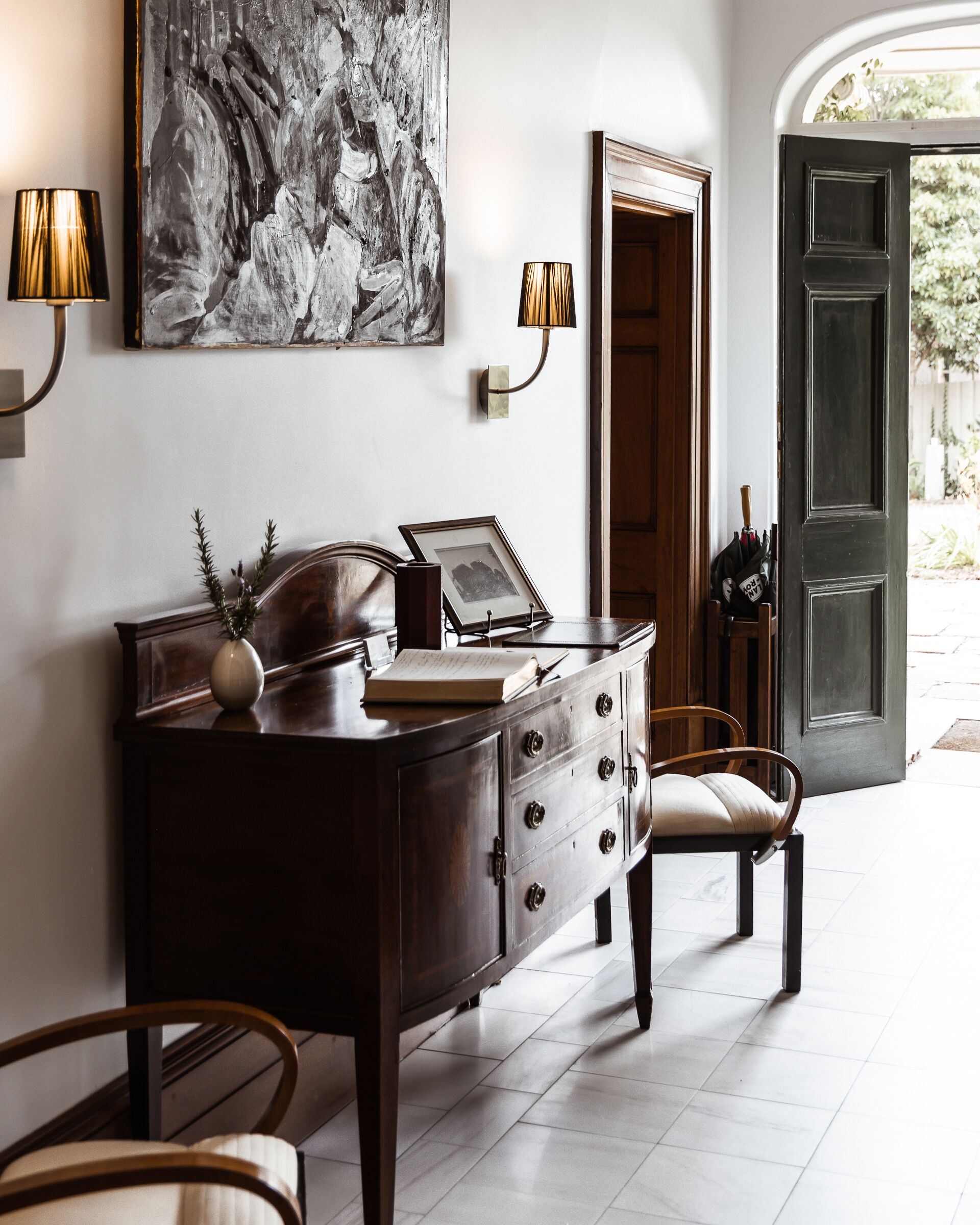 Un bureau en bois avec des tiroirs, un vase et un tableau encadré. Une chaise est placée devant le bureau. Une lampe est fixée au mur, à gauche du bureau.