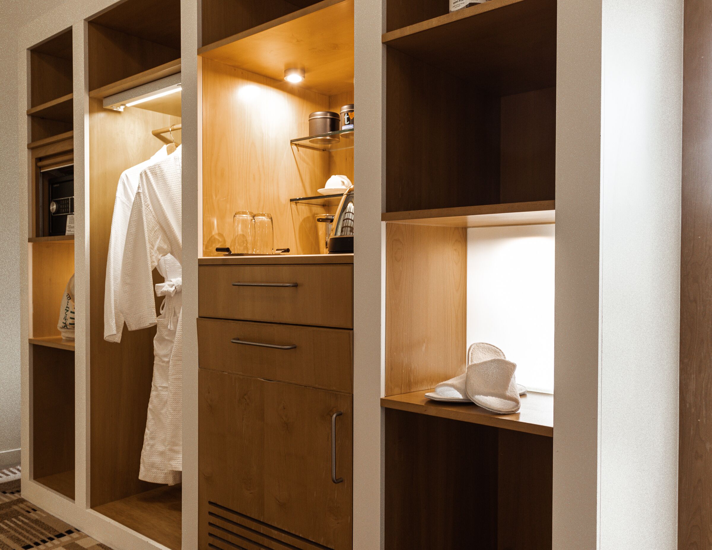 A wooden walk-in closet with a robe, a shelf with a lamp, and a pair of slippers.