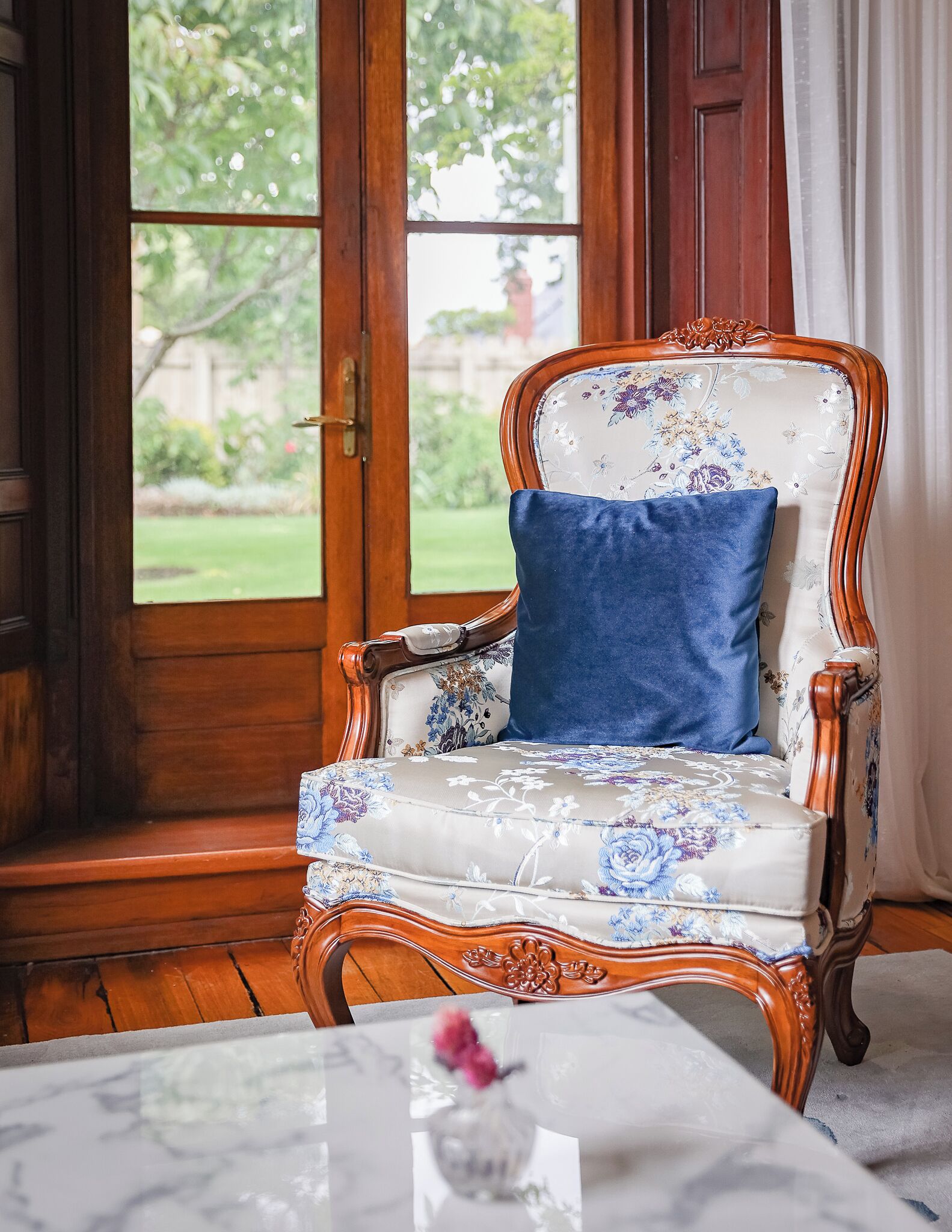 A chair with floral upholstery and a blue cushion, positioned near a window with a view of greenery.
