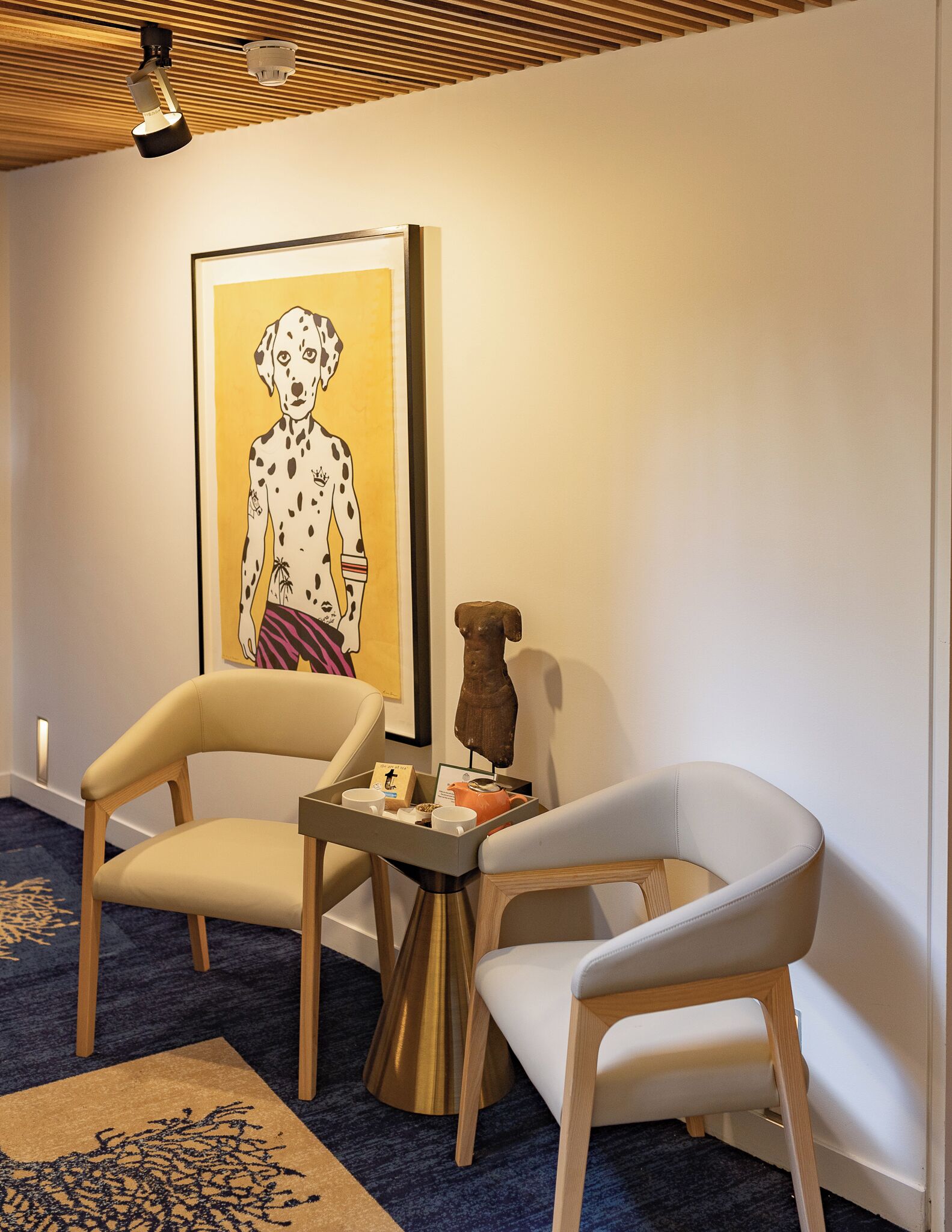A room with two white chairs, a small table with a tray, a framed artwork, and a wooden sculpture.